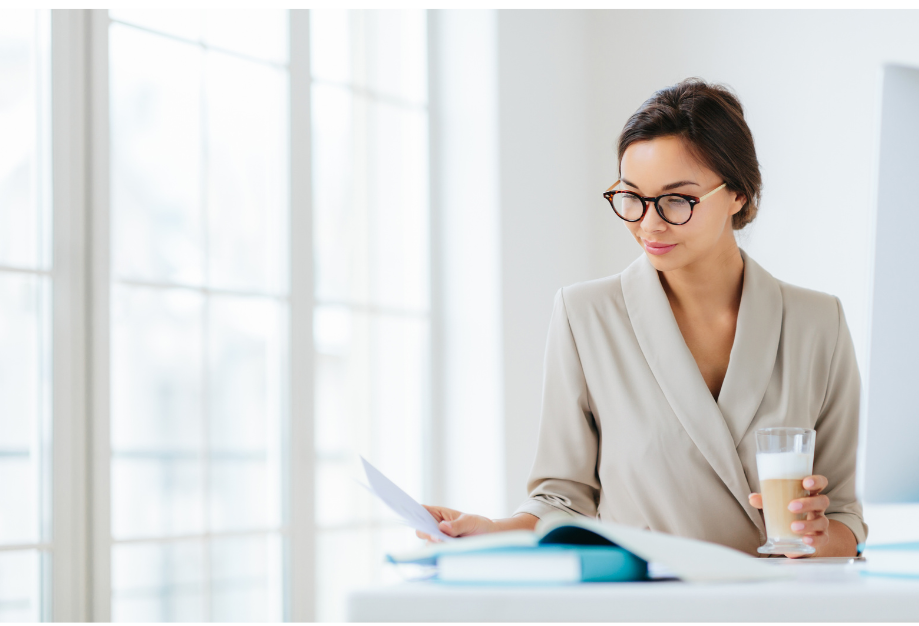 A career-driven woman in a tan business suit is reading at a table drinking a beverage.