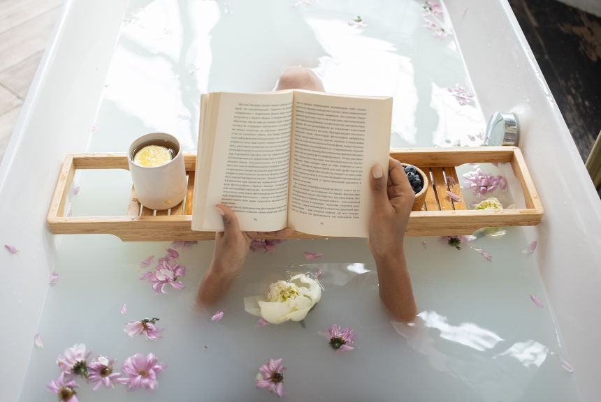 A woman is soaking and resting in a bathtub while reading a book.